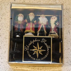 May include: A set of four Santa Claus figurines in a clear plastic box. Each figurine wears a red coat with white trim and holds a small green tree. The box has a gold border and a black label with the text "Christopher Radko HOME FOR THE HOLIDAYS".