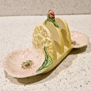 Op de afbeelding: Een keramische serveerschaal met bloementhema, met een lichtgeel midden en twee roze maïsvormige zijkanten. Het midden heeft drie verticale sleuven. De schaal is versierd met groene bladeren en een rode knop.