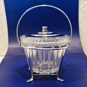 May include: A clear, faceted glass ice bucket with a silver-colored metal handle and trim. The bucket has a lid and sits on a silver-colored metal stand. The ice bucket is designed for serving ice.