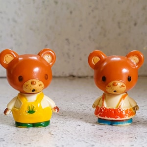 Può includere: Quattro figurine di orsi marroni con abiti diversi. L'orso a sinistra indossa un abito blu, l'orso successivo indossa un abito giallo e verde, il terzo orso indossa un vestito rosso e arancione e l'ultimo orso indossa un vestito bianco con una fascia rosa.