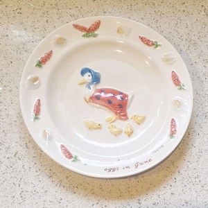 Puede incluir: Plato de cerámica blanca con un diseño de pato en relieve con un gorro azul y un chal a lunares rojos. El plato está decorado con zanahorias, conchas y el texto "more eggs in June".