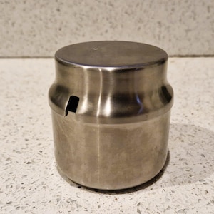 Peut inclure: Un récipient en acier inoxydable avec un corps cylindrique et un dessus plat et arrondi. Le récipient comporte une petite ouverture rectangulaire sur le côté. La surface du récipient a une finition en métal brossé.