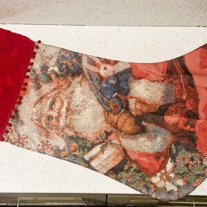 Puede incluir: Un calcetín navideño con una parte superior de terciopelo rojo y un cuerpo estampado con Papá Noel, regalos y vegetación festiva. El calcetín tiene un lazo para colgar y está adornado con pequeños pompones rojos.