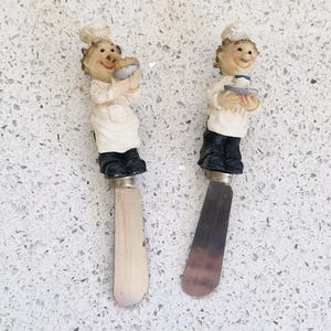 May include: Two decorative butter knives featuring chef figurines. The chefs wear white uniforms, black shoes, and chef hats. One holds a pie, the other a plate. The blades are silver and wood-toned.