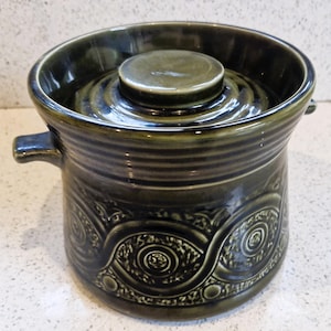 Könnte beinhalten: Ein dunkelgrüner Keramiktopf mit Deckel. Der Topf hat zwei kleine Griffe und ein dekoratives Muster am Boden. Der Deckel ist kreisförmig und passt gut. Der Topf ist etwa 15 cm hoch.