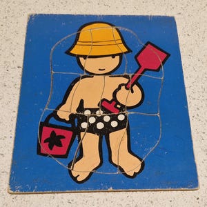 Puede incluir: Un rompecabezas de madera con un niño que lleva un sombrero amarillo y un traje de baño de lunares negros y blancos. El niño sostiene una pala roja y un cubo rojo con una estrella negra.