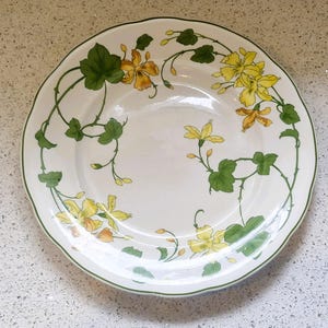Puede incluir: Plato de cerámica blanca con borde festoneado, adornado con un borde verde y un diseño floral. El diseño presenta flores amarillas y hojas verdes sobre vides sinuosas. Una pieza decorativa.