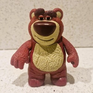 Peut inclure: Figurine d'ours rouge et beige avec un nez marron et un ventre jaune. L'ours a une expression s&eacute;v&egrave;re et se tient debout. Ce jouet de collection est issu de la franchise Toy Story.