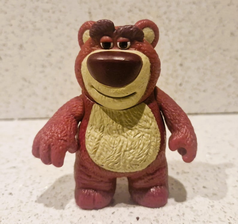 Peut inclure: Figurine d'ours rouge et beige avec un nez marron et un ventre jaune. L'ours a une expression s&eacute;v&egrave;re et se tient debout. Ce jouet de collection est issu de la franchise Toy Story.