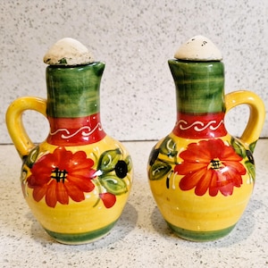 Può includere: Due oliere e acetai in ceramica. Ogni oliera ha una base gialla, una fascia rossa e bianca e un collo verde. Un grande fiore rosso con foglie verdi decora la parte anteriore. Ogni oliera ha un piccolo manico e un tappo beige.