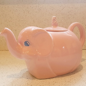 Puede incluir: Una tetera de cerámica rosa con forma de elefante con una pequeña figura de elefante en la tapa. La tetera tiene un pico y una asa.
