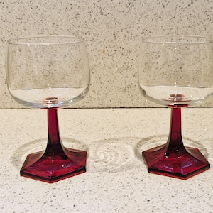 以下が含まれることがあります： 赤い六角形の台座が付いた透明なガラス製のワイングラスが2つ。グラスは空で、薄い灰色の表面の上に置かれています。
