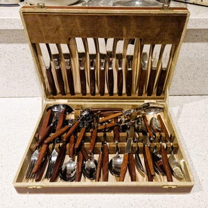 Puede incluir: Un juego de cubiertos de madera vintage en un estuche. El juego incluye cuchillos, tenedores, cucharas y utensilios para servir con hojas de acero inoxidable y mangos de madera marrón. El estuche está forrado con una tela beige.