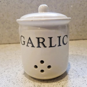 Może przedstawiać: Biały ceramiczny kubek z czarnym uchwytem i napisem "But First, Coffee" pogrubioną czcionką. Kubek jest ustawiony na rozmytym tle, sugerującym przytulne otoczenie.