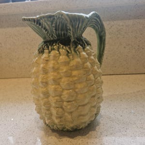 May include: A ceramic pitcher shaped like a pineapple. The pitcher has a textured, off-white body with a green leafy top and handle. The pitcher is a decorative item.