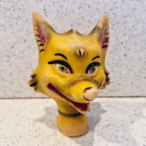 May include: A vintage, yellow fox head figurine with green eyes, black eyeliner, and a red mouth. The fox has pointed ears with pink inner details and a black collar. The figurine is made of a solid material.