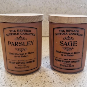 May include: Two terracotta-colored canisters with wooden lids. One is labeled "Parsley" and the other "Sage." Both have a vintage design with text that reads "The Revised Suffolk Canister" and "Henry Watson Pottery, England, Founded 1800."