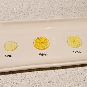 Może przedstawiać: Biała prostokątna tacka do serwowania z ilustracją plasterków limonki, pomarańczy i cytryny. Taca jest oznaczona jako "Lime", "Orange" i "Lemon".