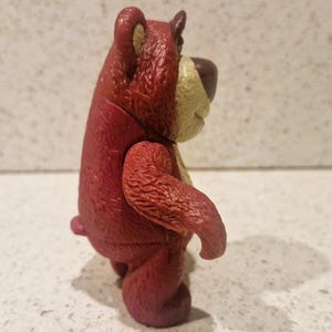 Peut inclure: Figurine d'ours en plastique rouge et beige. L'ours a une apparence textur&eacute;e, avec une couleur rouge plus fonc&eacute;e sur le corps et une couleur beige plus claire sur le museau et l'int&eacute;rieur des oreilles. L'ours est debout, les bras le long du corps.
