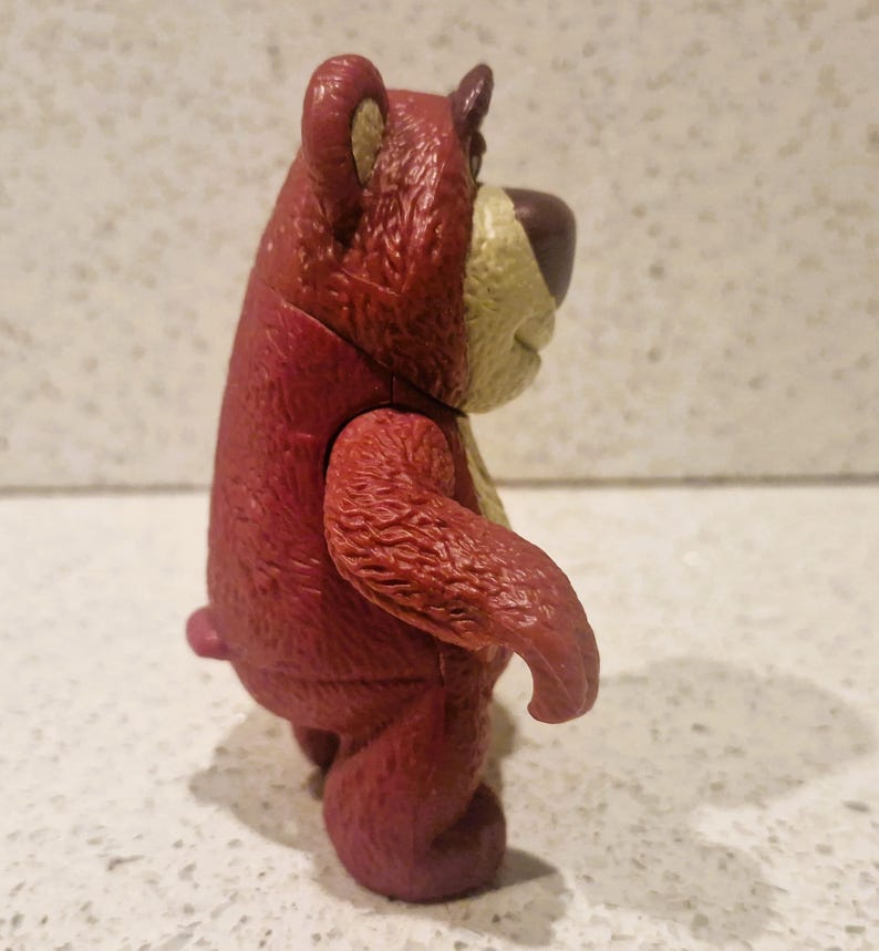 Peut inclure: Figurine d'ours en plastique rouge et beige. L'ours a une apparence textur&eacute;e, avec une couleur rouge plus fonc&eacute;e sur le corps et une couleur beige plus claire sur le museau et l'int&eacute;rieur des oreilles. L'ours est debout, les bras le long du corps.