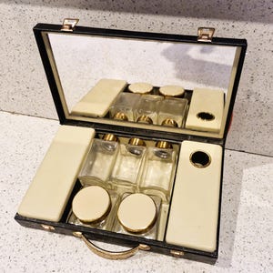 Puede incluir: Un neceser negro vintage abierto con espejo, con frascos y tarros de vidrio con tapas doradas. El estuche incluye compartimentos de color marfil y un asa dorada. El estuche mide aproximadamente 25 cm de ancho.
