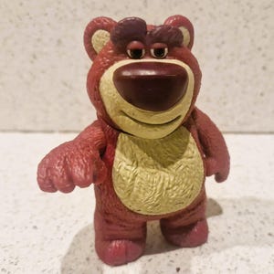 Peut inclure: Un ours en plastique rouge et beige avec un nez brun fonc&eacute; et une expression souriante. L'ours a un ventre beige clair et se tient debout, les bras tendus. Le jouet est un personnage du film d'animation Toy Story.
