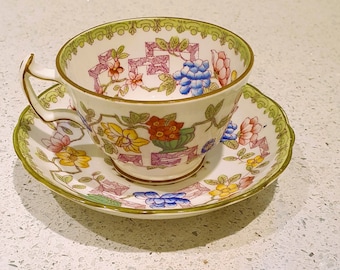 Può includere: Un set vintage di tazza e piattino da tè con un motivo floreale in tonalità di rosa, giallo, blu e verde. La tazza e il piattino hanno un bordo dorato e un bordo verde.
