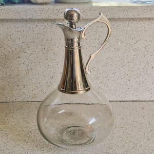 May include: A clear glass decanter with a silver-plated top and handle. The decanter has a round body and a long, slender neck.