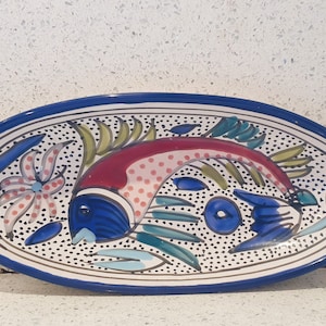 Op de afbeelding: Een ovale serveerschaal van keramiek met een blauwe rand en een witte achtergrond. De schaal heeft een kleurrijk handgeschilderd ontwerp van een vis met rode, blauwe, groene en gele accenten. De vis is omgeven door een witte achtergrond met kleine zwarte stippen.