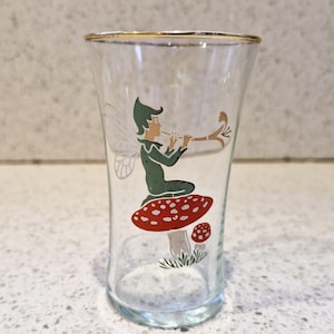 May include: A clear glass shot glass with a gold rim. The glass features a whimsical illustration of a fairy playing a flute while sitting on a red and white mushroom.