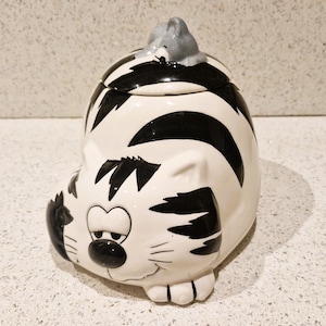 Può includere: Un barattolo per biscotti a forma di gatto in ceramica, bianco con strisce nere. Il barattolo ha una faccia da cartone animato con tratti neri e baffi. Una piccola statuina di gatto grigio è sul coperchio.