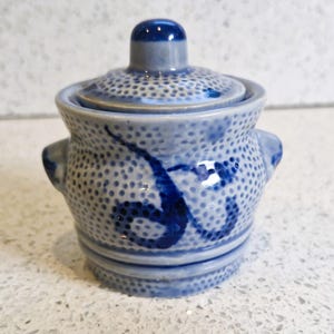 以下が含まれることがあります： 蓋付きの小さな青と白のセラミックジャー。ジャーは、テクスチャード加工されたドットパターンと濃い青色の抽象的なデザインが特徴です。2つの小さなハンドルと、ノブが付いた丸い蓋が付いています。