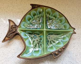 May include: A green and blue ceramic fish-shaped serving dish with four sections. The dish has a brown rim and a seahorse design on the tail.