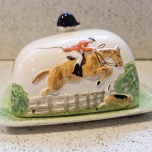 Peut inclure: Un beurrier en céramique avec un couvercle représentant un cheval et un cavalier sautant une barrière. Le couvercle a un bouton noir. La base est vert clair.