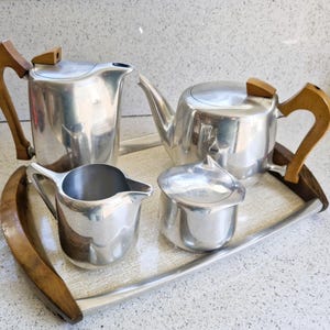 May include: A vintage tea set with a teapot, creamer, sugar bowl, and a serving pitcher. The set is made of polished silver metal with wooden handles and accents. The set is presented on a rectangular tray with wooden edges.