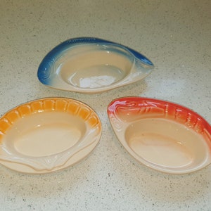 Op de afbeelding: Drie keramische kommen in de vorm van vissen. De kommen zijn in tinten blauw, oranje en rood. De kommen zijn allemaal leeg.