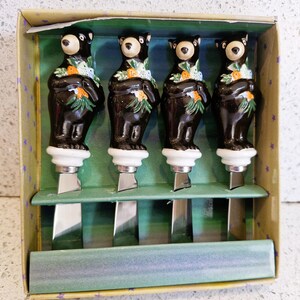 Peut inclure: Un ensemble de quatre couteaux à fromage dans une boîte décorative. Chaque couteau est orné d'un manche en forme d'ours noir avec une base blanche et une lame en acier inoxydable. Les ours ont des motifs floraux.