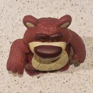 Peut inclure: Gros plan d'une figurine d'ours brun-rouge avec un nez brun fonc&eacute; et les yeux ferm&eacute;s. L'ours a un ventre beige clair et est en position assise. La figurine est un jouet.