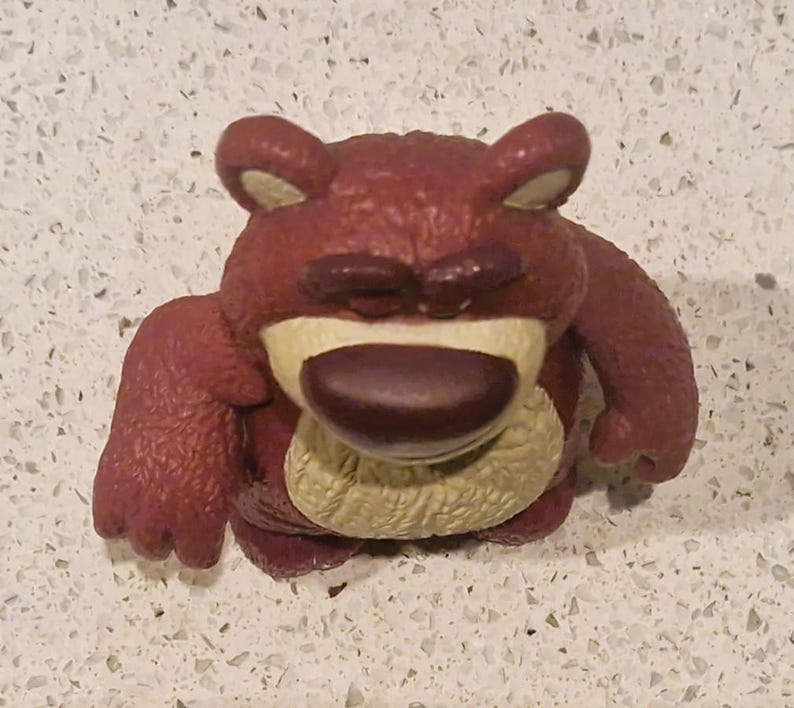 Peut inclure: Gros plan d'une figurine d'ours brun-rouge avec un nez brun fonc&eacute; et les yeux ferm&eacute;s. L'ours a un ventre beige clair et est en position assise. La figurine est un jouet.