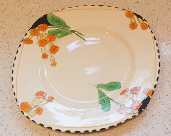 May include: A white ceramic plate with a black and white dotted border. The plate features a floral design with orange and yellow flowers and green leaves.