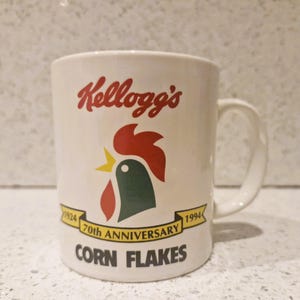 Op de afbeelding: Witte keramische mok met het Kellogg's-logo in rood schrift en een haan-afbeelding in rood, groen en geel. De mok viert de 70e verjaardag van Corn Flakes, met tekst en de data 1924-1994.