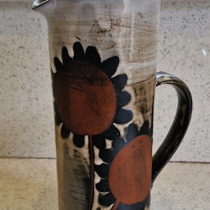 Op de afbeelding: Hoge keramische kan met deksel, versierd met een zonnebloemontwerp in bruin, zwart en oranje. De kan heeft een handvat en een schenktuit. Het ontwerp is rustiek en handgemaakt, geschikt voor het serveren van dranken. Ongeveer 25 cm hoog.