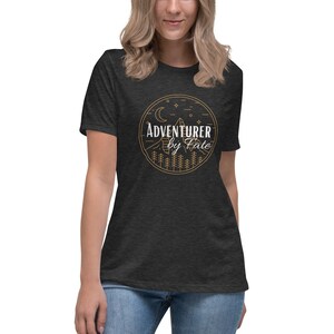 以下が含まれることがあります： 暗いヘザーグレーのTシャツに、円の中に山脈、月、星が描かれた金色のグラフィックがプリントされています。円の中に「Adventurer by Fate」という文字が白で書かれています。