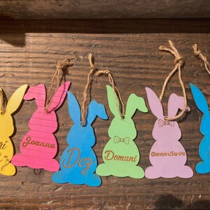 May include: Six wooden bunny shaped ornaments with names written on them. The bunnies are painted in different colors: yellow, pink, blue, green, purple, and blue. Each bunny has a twine string for hanging.
