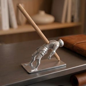 May include: A silver-colored metal knight figurine pen holder mounted on a rectangular base. The knight is depicted in a reclining position, holding a light-colored wooden pencil diagonally in its hands, which serves as a functional desk accessory.