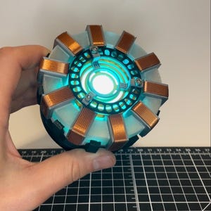 Op de afbeelding: Een cirkelvormig arc-reactor rekwisiet met een gloeiend cyaan licht in het midden, omringd door tien koperkleurige draadspoelen die radiaal zijn geplaatst. Het apparaat heeft een wit doorschijnend frame met zilverkleurige accenten en schroeven.