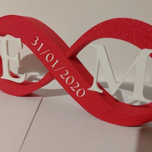 Può includere: Un simbolo dell'infinito rosso con dettagli glitterati bianchi, con le lettere "E" e "M" e la data "31/01/2020". Le lettere sono bianche. Un oggetto decorativo.