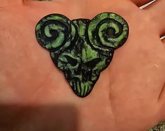 Può includere: Un plettro per chitarra triangolare con un design nero e verde neon. La superficie presenta un teschio stilizzato con prominenti corna di ariete a spirale sulla parte superiore, in una finitura testurizzata ad alto contrasto.