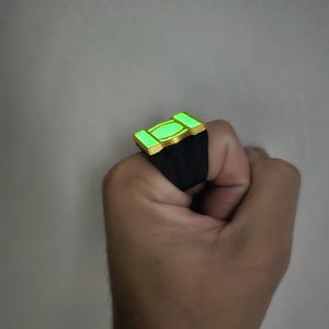 Op de afbeelding: Een ring met een zwarte band en een rechthoekige goudkleurige zetting met een oplichtend lichtgroen middenpaneel. Het ontwerp bevat een centrale ovale vorm geflankeerd door twee kleinere rechthoekige secties die een fel neon-groen licht uitstralen.