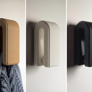 Op de afbeelding: Drie wandhaken in beige, wit en zwart. Elke haak heeft een U-vormig ontwerp met verticale ribbels op het buitenoppervlak. Een grijze, gestructureerde handdoek hangt aan de beige haak, terwijl de andere twee haken leeg zijn.